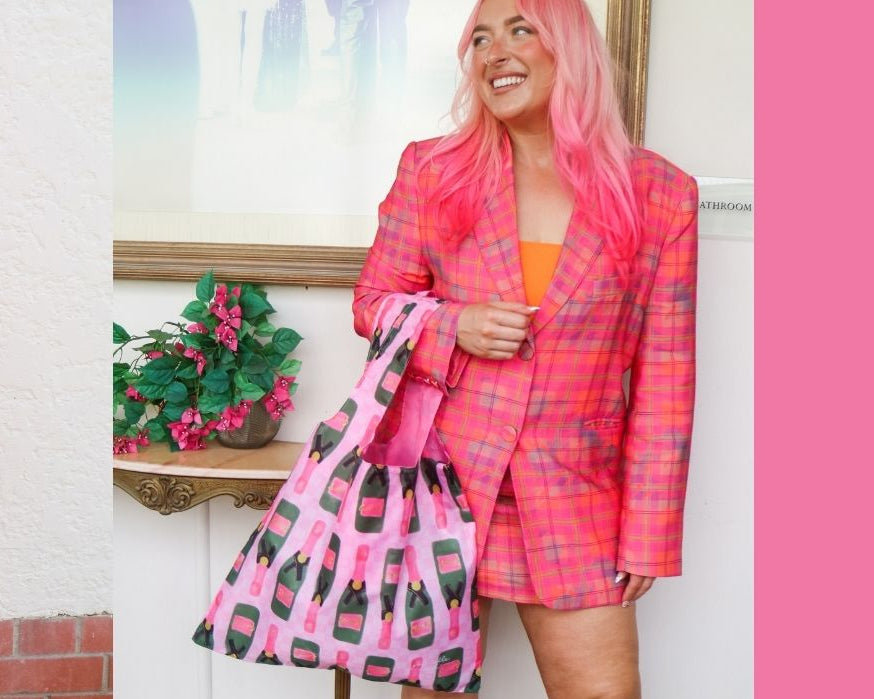 A split-screen lifestyle image showing a woman with pink hair wearing a pink plaid blazer and holding a pink shopping bag with 'Girl Dinner' written on it. The left side shows her outdoors against a white wall, and the right side shows her indoors against a framed picture, with a large solid pink block on the right side of the image.