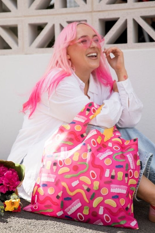 A pink-themed lifestyle collage featuring a model with bright pink hair wearing pink-tinted sunglasses and a white shirt, sitting on a stone ledge next to a large pink and multicolor sprinkle-patterned tote bag. The background is a clean white wall with a geometric pattern, and a solid bright pink block is on the right side.