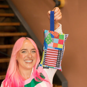A woman with bright pink hair holds up a small square patchwork fabric pouch with a blue handle. The pouch features a colorful patchwork design with red, green, yellow, blue, and black patterned sections.