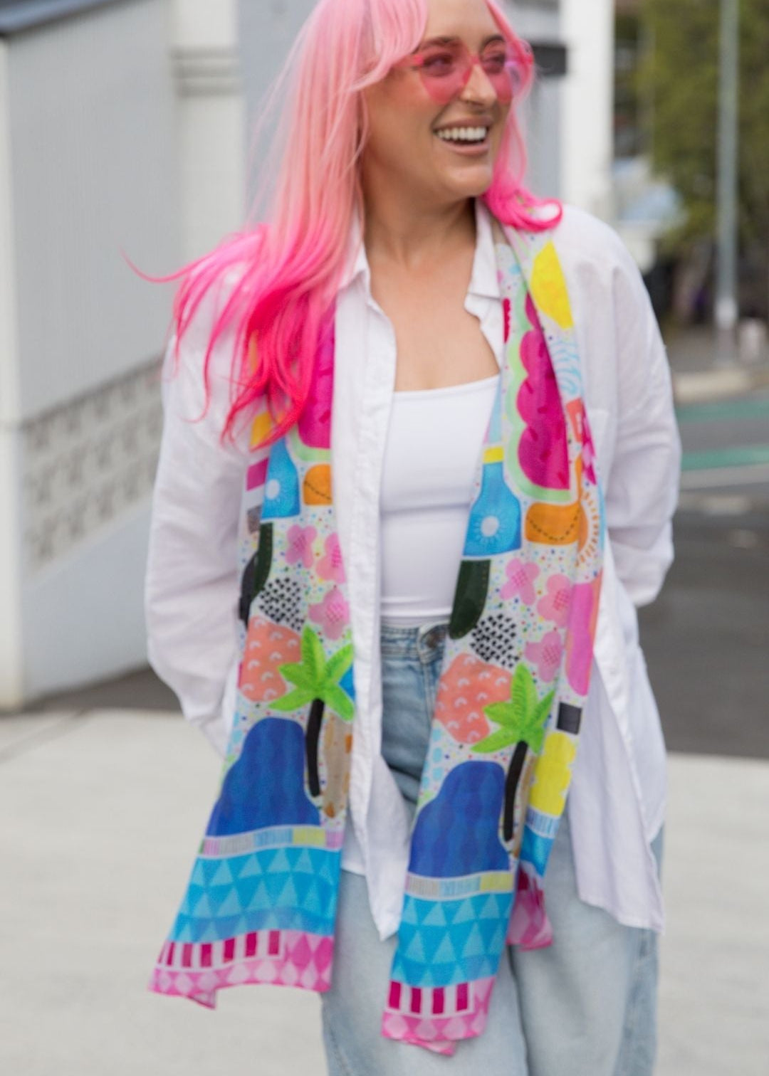 A woman with bright pink hair and pink-tinted sunglasses is smiling outdoors, wearing a white button-up shirt, light blue jeans, and a colorful abstract-patterned scarf.
